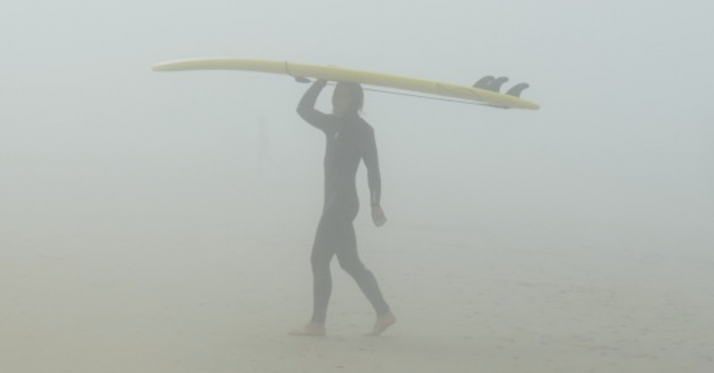 Zdjęcie przedstawia człowieka z deską surfingową w szarej mgle.
